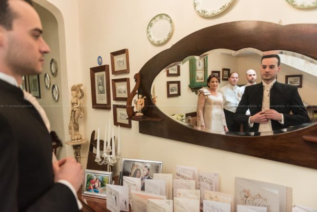 groom getting ready in front of the mirror, with his family observing him from behind