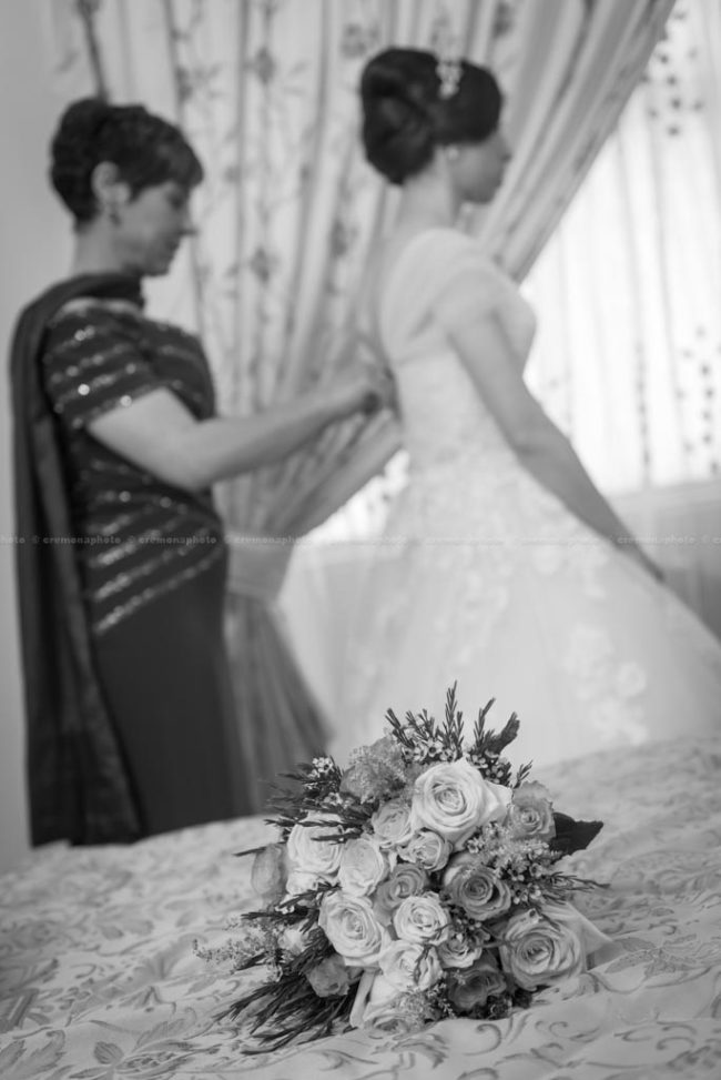 A flower bouquet is laying on the bed, with mother helping her daughter into the bridal dress