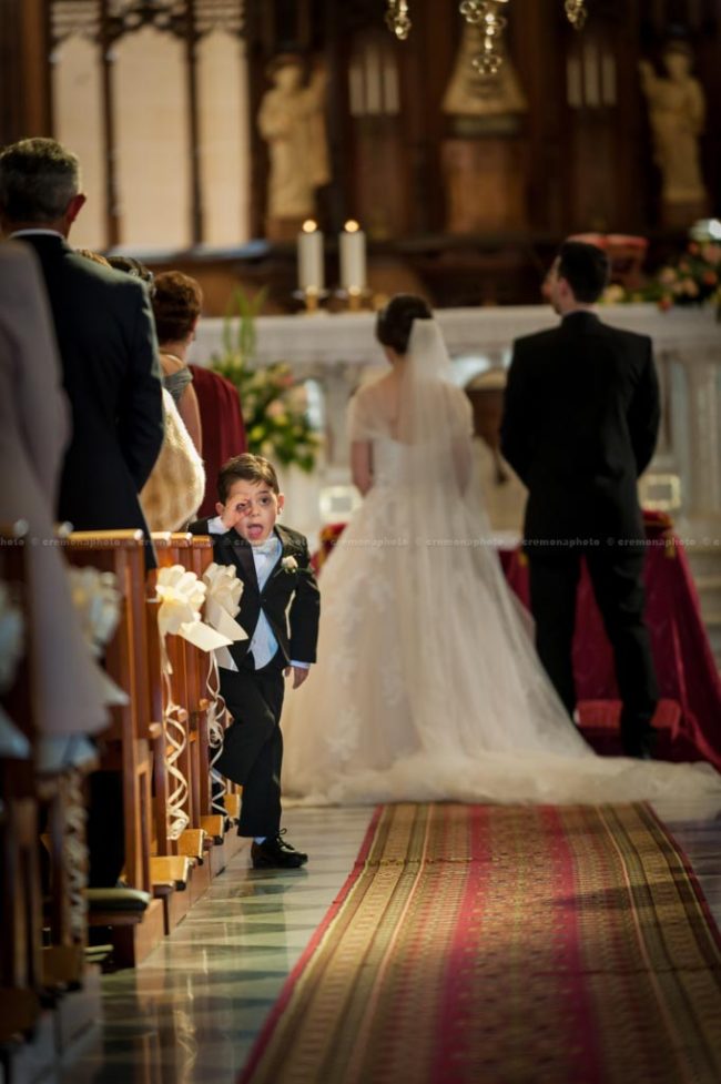 A young page boy pulling up a silly face, behind the back of the bride and groom