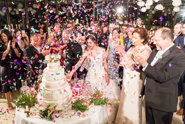 Bride and groom cutting the wedding cake surrounded by family, friends and a rainfall of colorful paper confetti